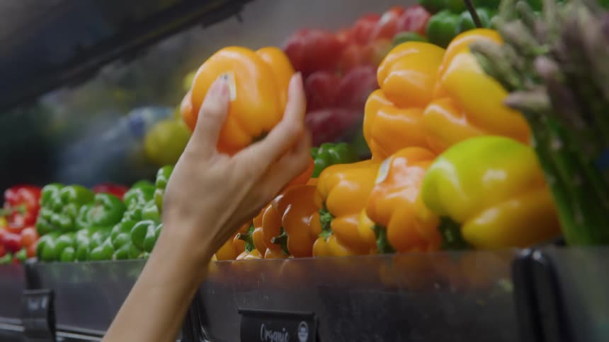 Woman reaches for a pepper in a grocery store and takes it from the shelf