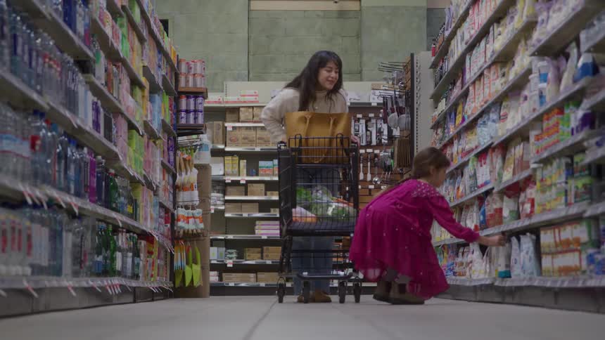 A woman pushing a shopping cart down a supermarket aisle and pointing to a product for her little daughter to pick up