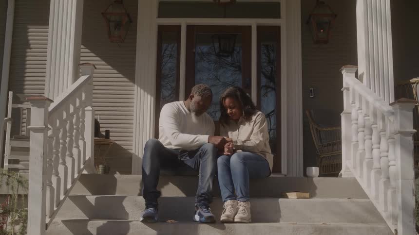 Two people seated on house steps share a reflective moment with clasped hands