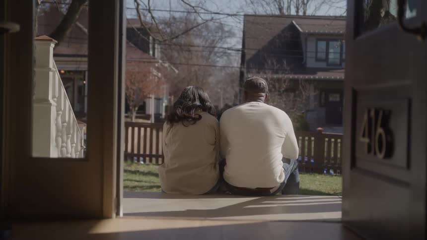 A man and a woman are sitting sidebyside on their front porch steps with their backs to the camera framed by the open doorway as they look out onto a quiet sunlit residential street with houses and leafless trees
