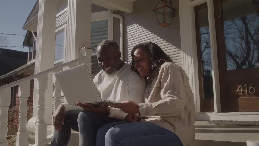 Two people sitting on front steps wave at laptop outside a house