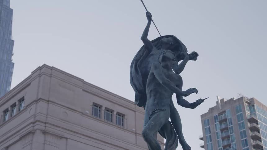 A dramatic bronze sculpture featuring two muscular figures one holding a spear aloft is framed against a bright clear sky contrasting with the beige classical architecture and modern skyscrapers surrounding it
