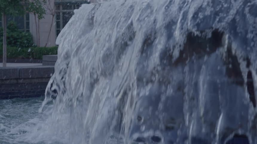 A large volume of water continuously pours over the rough curved surface of an outdoor fountain creating a thick churning white cascade that falls into a basin below set against a blurry urban background