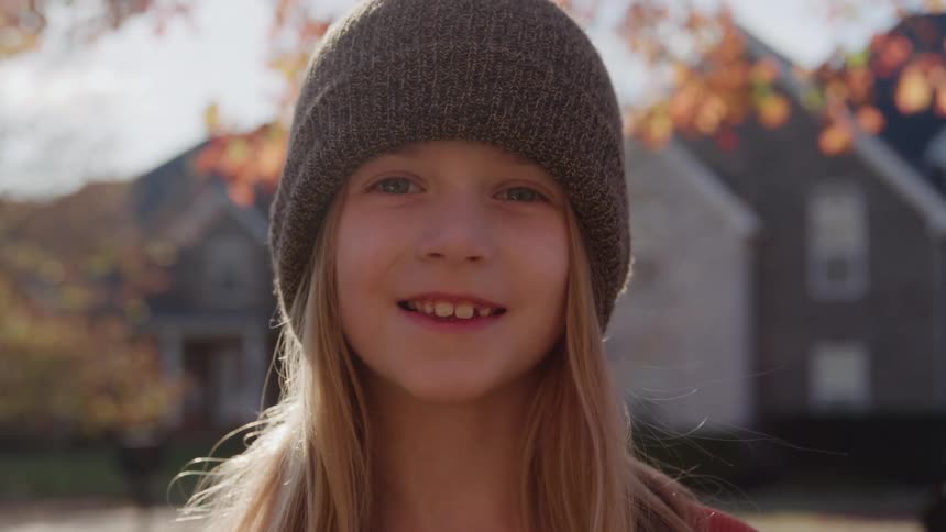 A medium closeup captures a young girl with long blonde hair and a dark beanie who is smiling broadly at the camera while standing outdoors in a bright suburban neighborhood during the autumn season