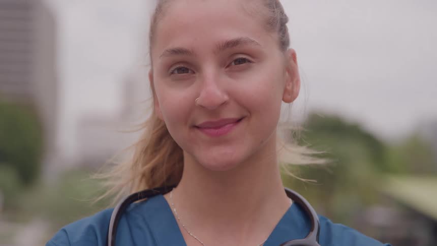 A young woman wearing blue medical scrubs and a stethoscope turns from a side view to face the camera with a confident gentle smile against a blurred bright backdrop of city buildings