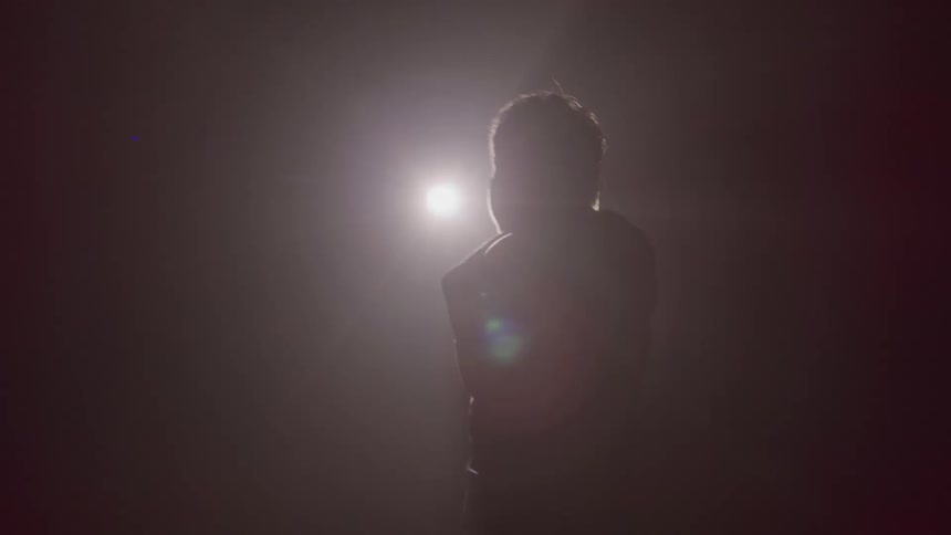 A person in a dark setting is silhouetted by a bright circular light source behind them briefly turning to show their profile waves and bows before leaving