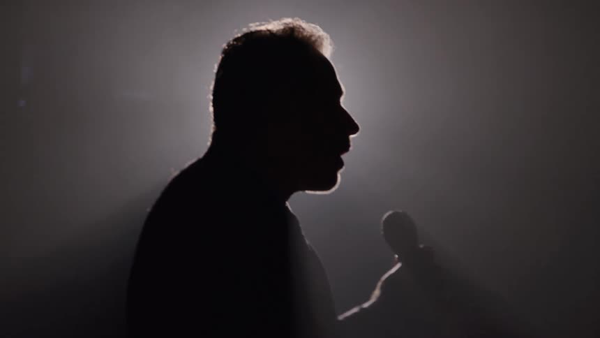 A side profile view shows the silhouette of a man standing on a stage speaking or singing into a handheld microphone with his figure heavily backlit by a powerful hazy light source
