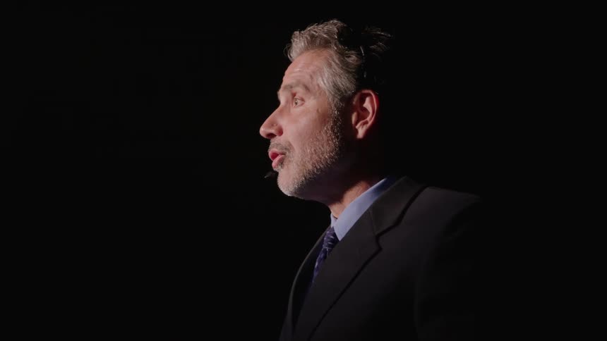 Man in suit gestures while speaking under dramatic lighting
