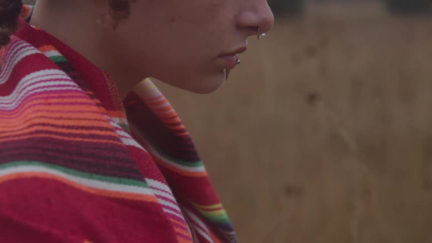 Profile of a young person with piercings wrapped in a colorful shawl in front of a dry field
