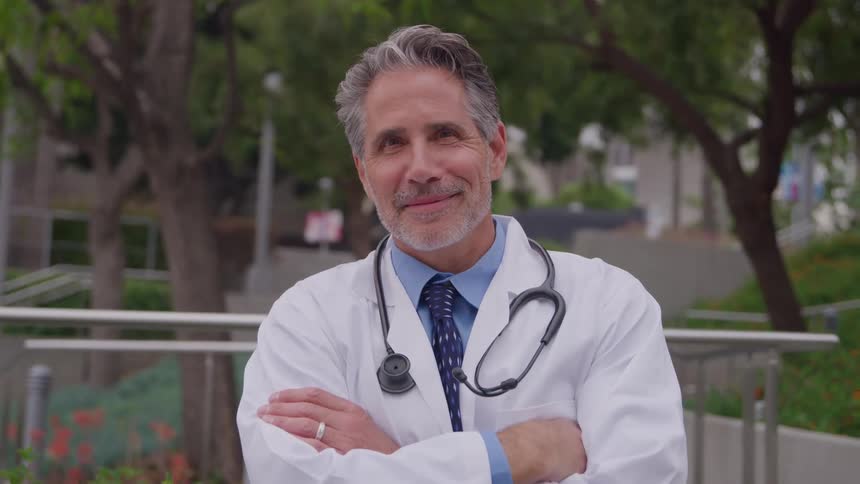 Doctor in a lab coat with a stethoscope smiles with arms crossed outdoors