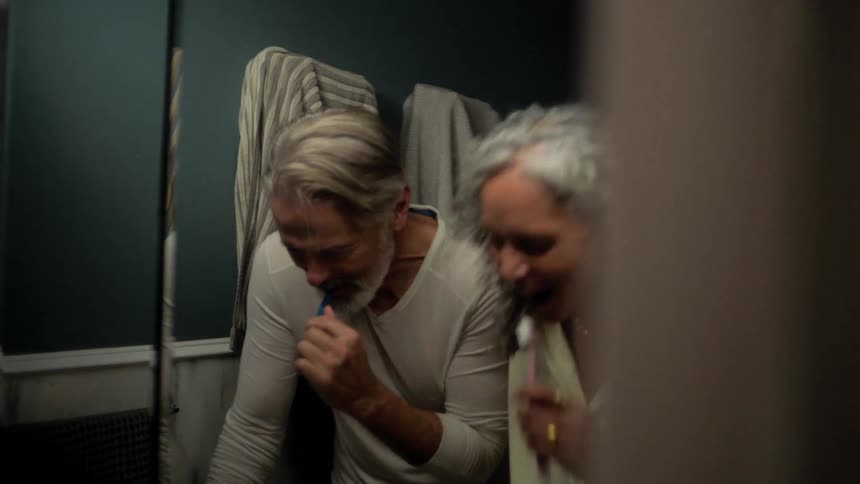 Couple smiles while brushing their teeth side by side in front of the mirror