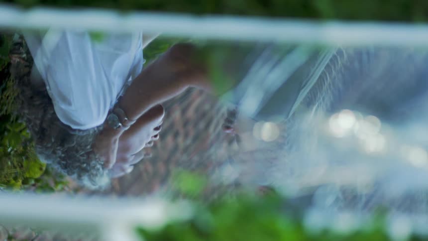 A person with curly graying hair wearing a white shirt and a watch is seen in an upsidedown reflection in a surface that appears to be water or wet glass which distorts the image and showsanother person holding the lady over a patch of grass