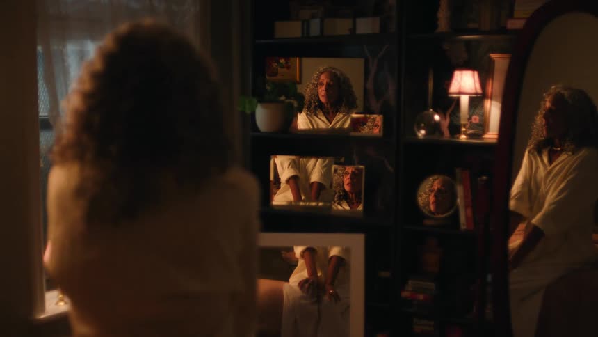 A woman with curly hair is reflected in multiple mirrors and framed photos across a dark richly decorated bookshelf in a bedroomthe reflection in a floor mirror shows her sitting down while the other mirrors and frames create a complex repeated ima