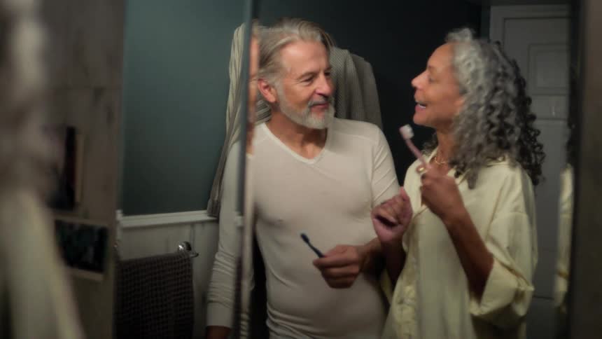 A man and woman are standing in a bathroom brushing their teeth talking and smiling while wearing pajamas