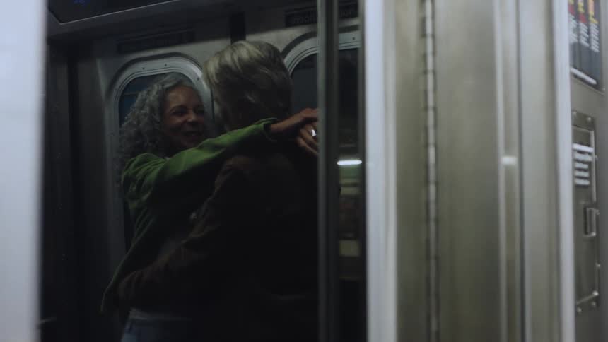 An older couple are standing and hugging on a moving subway at night
