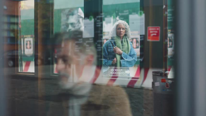 Woman looks through the storefront window as a soft reflection of another person passes across the scene