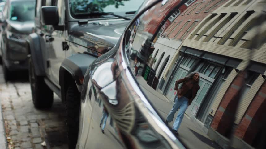 Reflections of a brick building slide across a glossy black car door as a silverhaired woman walks by her distorted image moving smoothly over the dark metallic surface