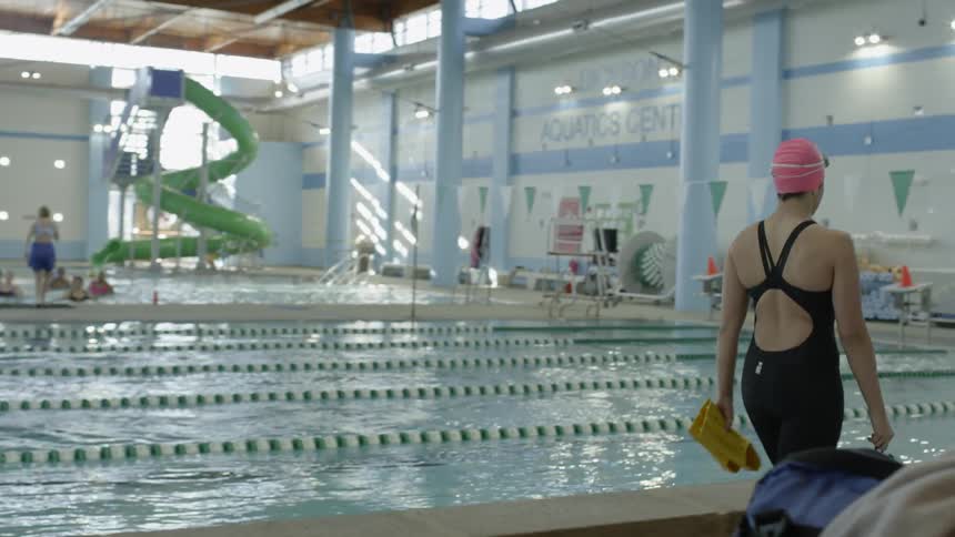 A swimmer walks by a large indoor lap pool carrying flippers