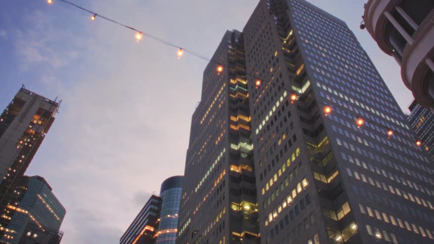 A series of highrise and skyscraper buildings stand tall in the city at midday their glass facades gleaming as sundown approaches streams of light shine out from the windows casting a warm glow across the skyline