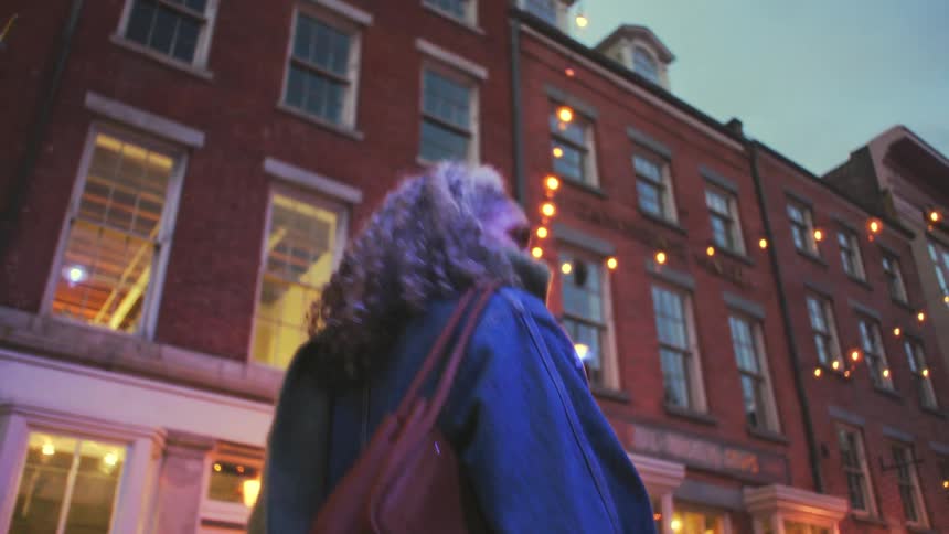 A person with bright curly hair walks along a city street at dusk looking upward toward the multistory red brick buildings the scene is illuminated by warm string lights and glowing windows creating a cozy evening atmosphere