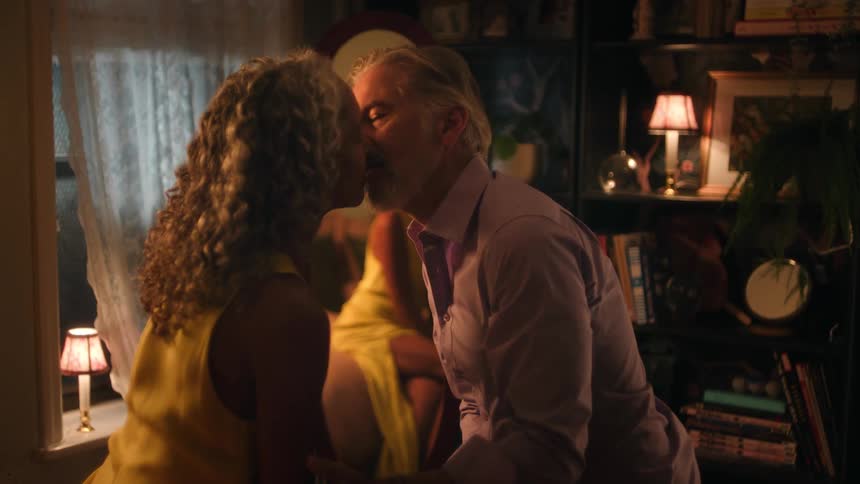 A man and a woman are standing close together in a dimly lit densely decorated room sharing an intimate kiss before separating with the womans seated reflection prominently visible in a large mirror behind them