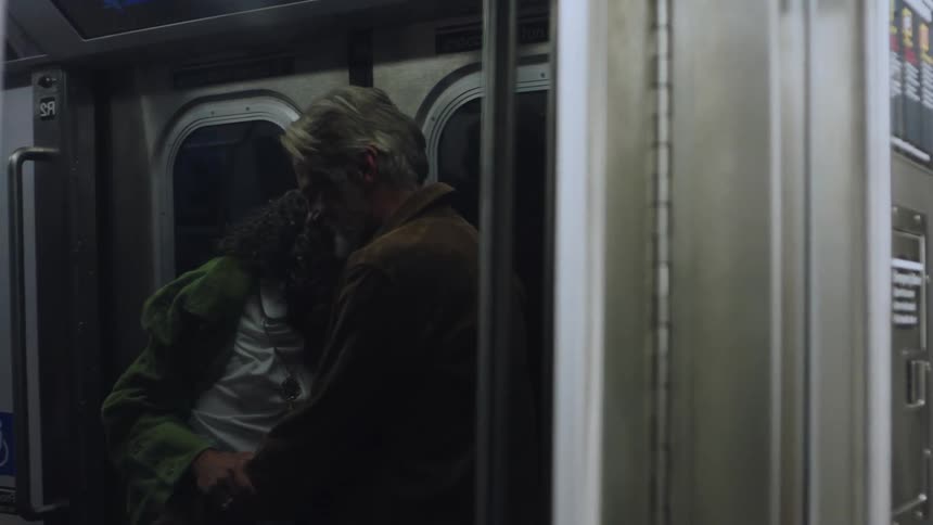 Inside a subway car a grayhaired woman with a joyful expression wearing a white shirt with a pendant necklace and a green jacket is warmly embracing a lighthaired person whose back is partially turned away