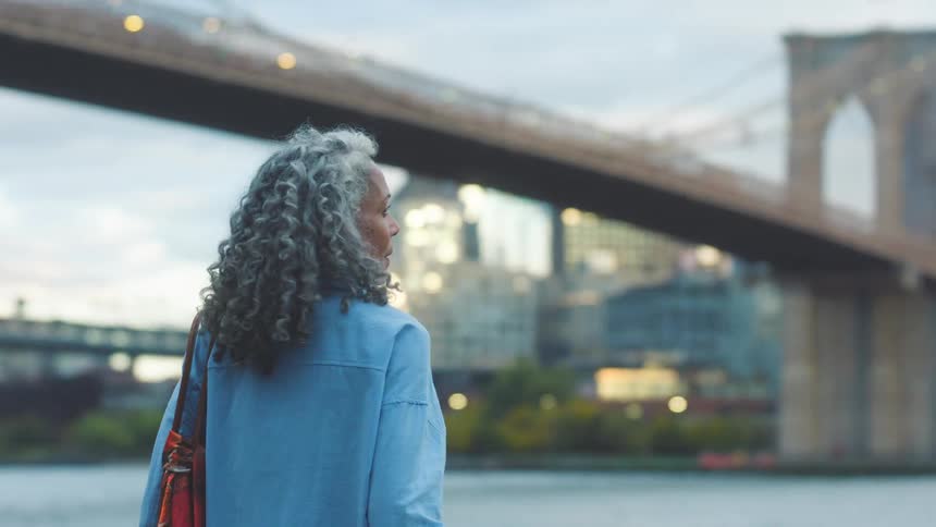 A woman leans on a railing by the waterfront while looking out toward a city bridge