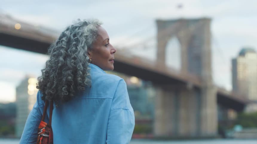 An older woman with long curly gray hair dressed in a blue shirt and jeans is walking on an urban waterfront before stopping to lean on a railing looking out with a bridge at the distance