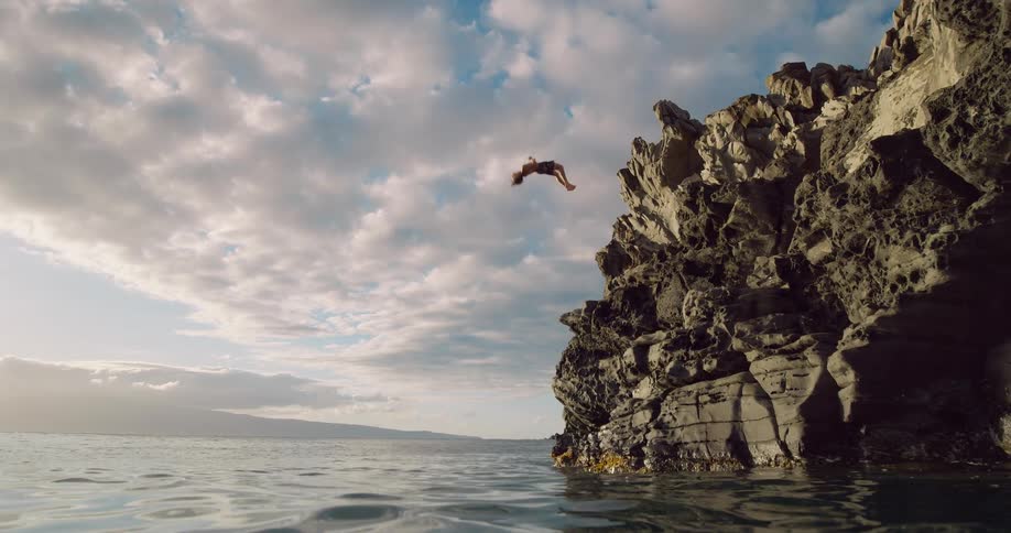 A person stands at the top of a rugged dark cliff jutting out of the ocean against a dramatic cloudy skythe person dives headfirst off the cliff and plunges into the water below creating a large splash