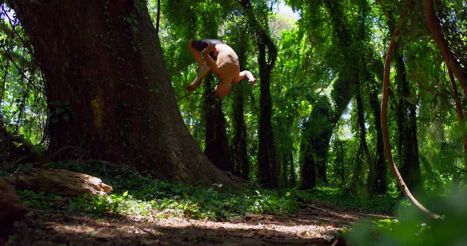 A shirtless man is performing a backflip in a lush forest