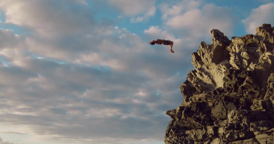 Man backflips off a seaside cliff while diving into the water on a cloudy day