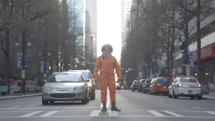 A man in an orange spacesuit and helmet stands in a crosswalk