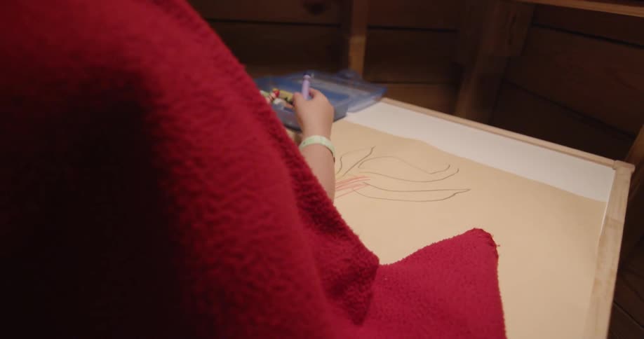A child wearing a blanket draws with crayons on paper