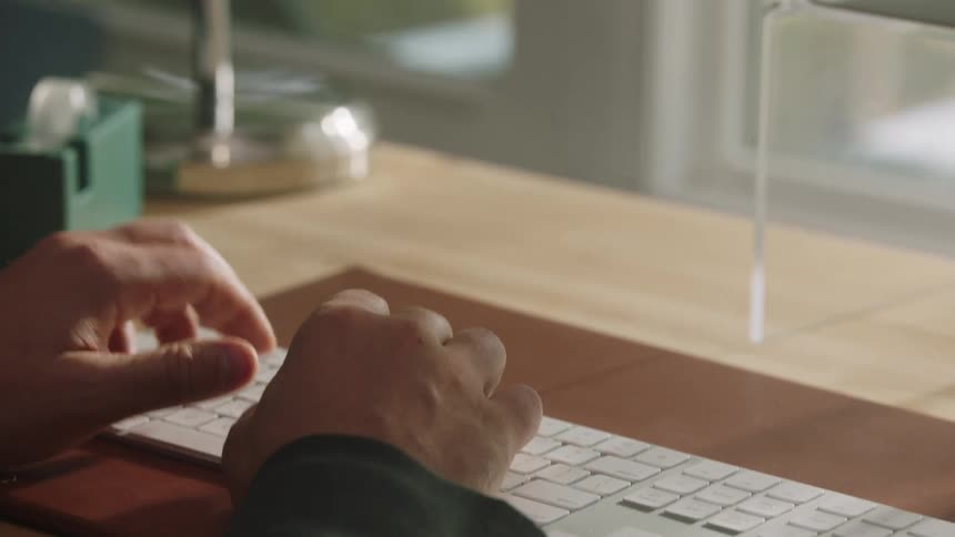 A closeup view from above shows a persons hands positioned over the keys of a white computer keyboard which rests on a wooden desk with a brown pad while in the background a window provides natural light and various desktop items are visible