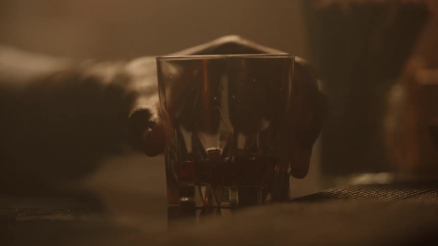 A hand quickly enters the frame to set a heavy faceted drinking glass containing a small amount of amber liquid down onto a dark surface in a warmly lit hazy environment