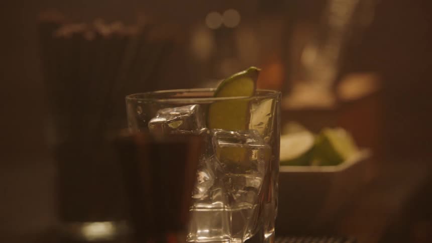 A bartender rubs a lime along the rim of an alcohol glass then drops the lime into the drink filled with ice as the scene unfolds in the bar someone reaches forward to pick up the glass