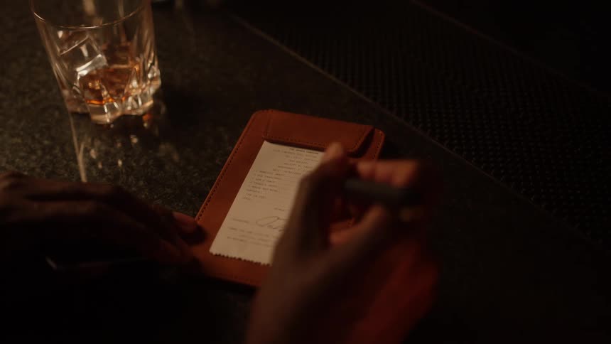 A customer at a bar retrieves their credit card from a bill holder and signs the receipt next to a glass of whiskey