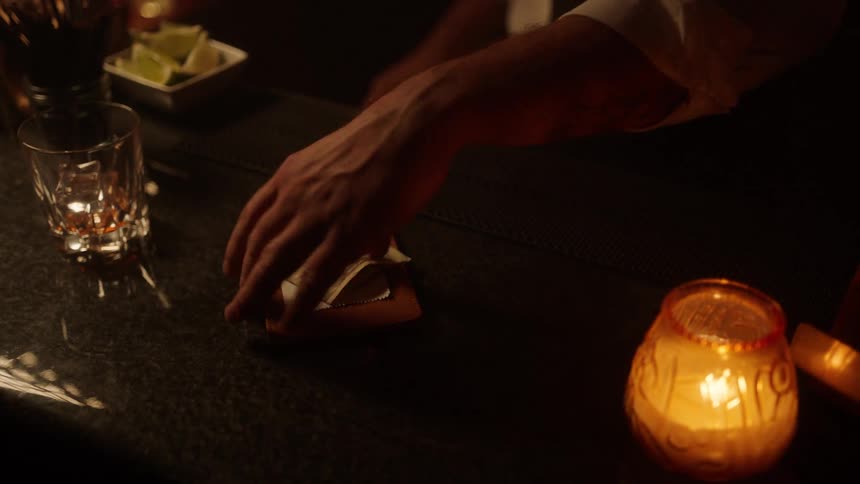 A bartender drops a tab on the counter as a customer pays cash for a drink an empty glass rests nearby with a candle glowing in a glass jar while the bartender picks up both the cash and the tab