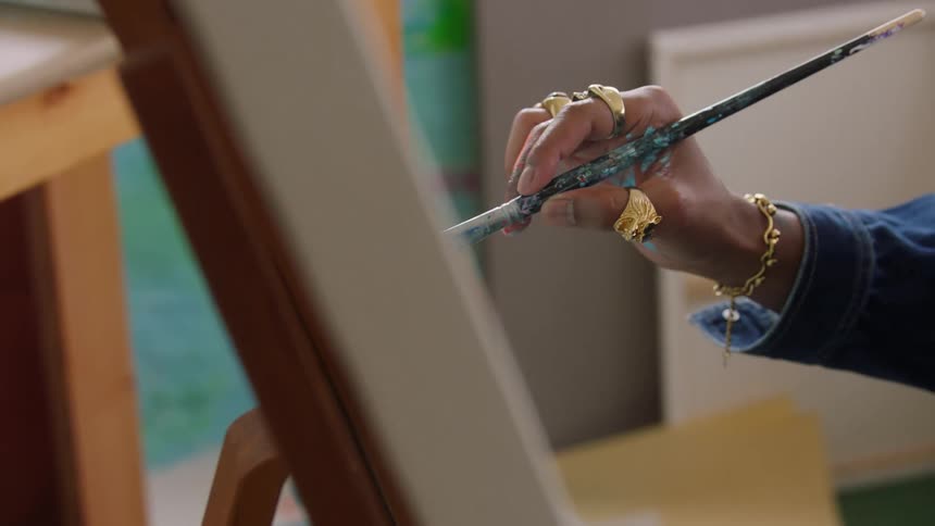The video is a closeup of an artists hand adorned with several gold rings and a bracelet the artist is holding a paintbrush and applying paint to a canvas resting on a wooden easel
