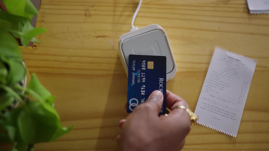 A closeup overhead view shows a persons hand holding a blue payment card and tapping it briefly onto a small white electronic card reader that is resting on a wood grain countertop next to a paper receipt