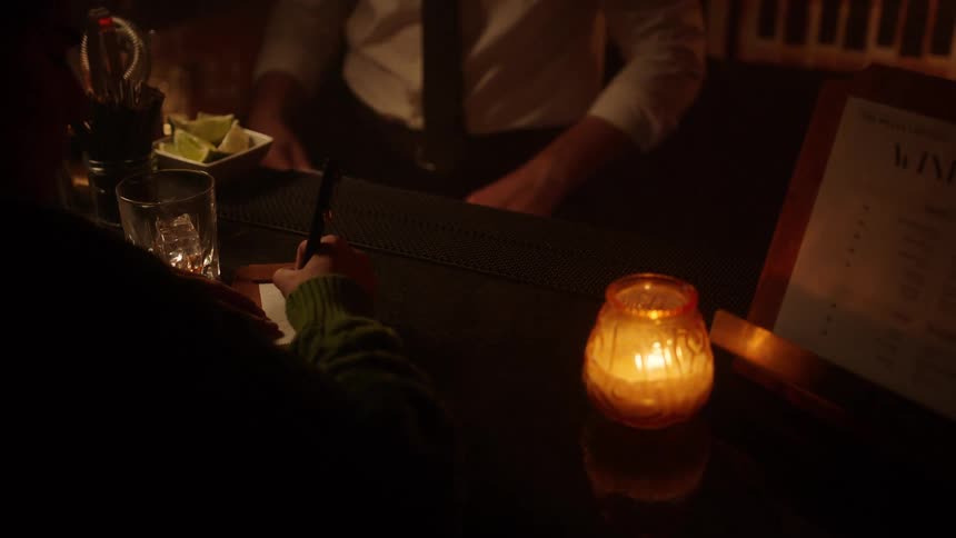 A bartender dressed in a white shirt and tie places a bill holder on the dark bar counter for a customer who signs the receipt illuminated by the glow of a nearby candle in a cozy dark bar setting