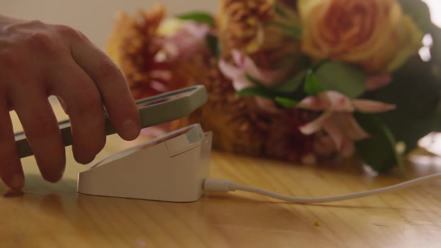 A persons hand wearing a silver digital wristwatch holds a small gray payment device and moves it to connect with a white wired pointofsale terminal on a wooden counter next to a blurry bouquet of flowers