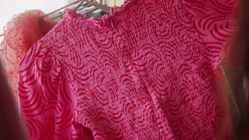 A persons hand reaches in to feel and then pull a pink and red smocked top or dress off of a clothing hanger while browsing a rack of various garments