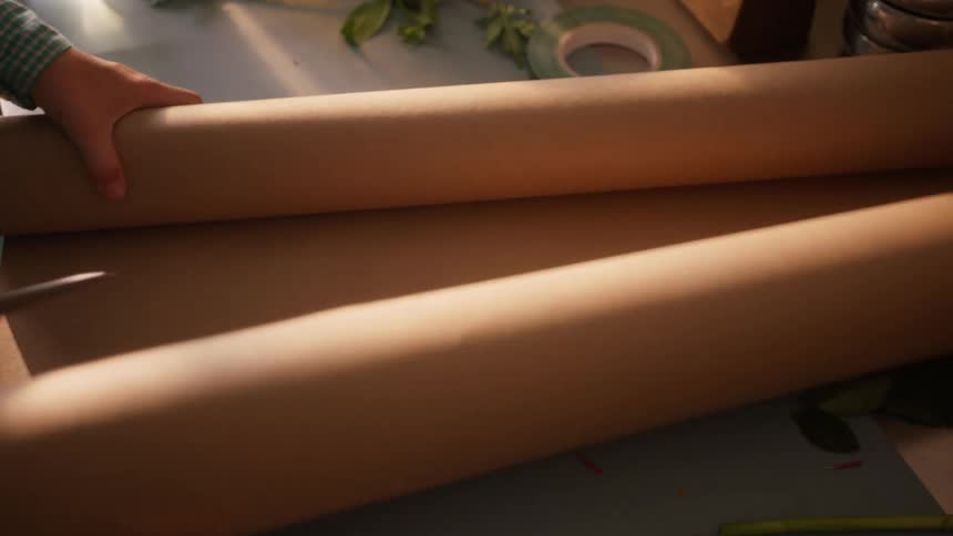 A closeup view focuses on a work surface where a person is unrolling a section from a large roll of brown wrapping paper flattening it against the counter and then using scissors to cut the sheet in a setting surrounded by crafting materials