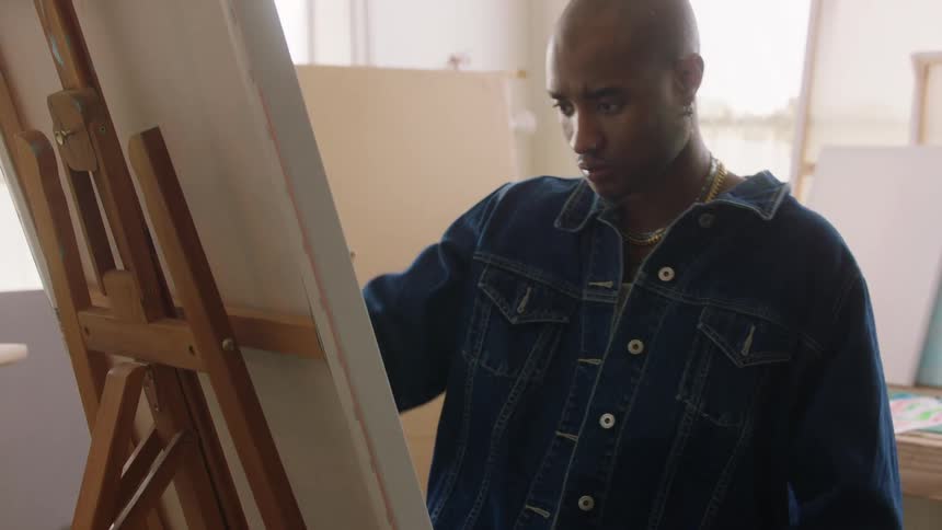An artist wearing a denim jacket and jeans sitting in a brightly lit studio in front of a canvas on a wooden easel holding a small brush and concentrating on the work while moving his arm away from the painting
