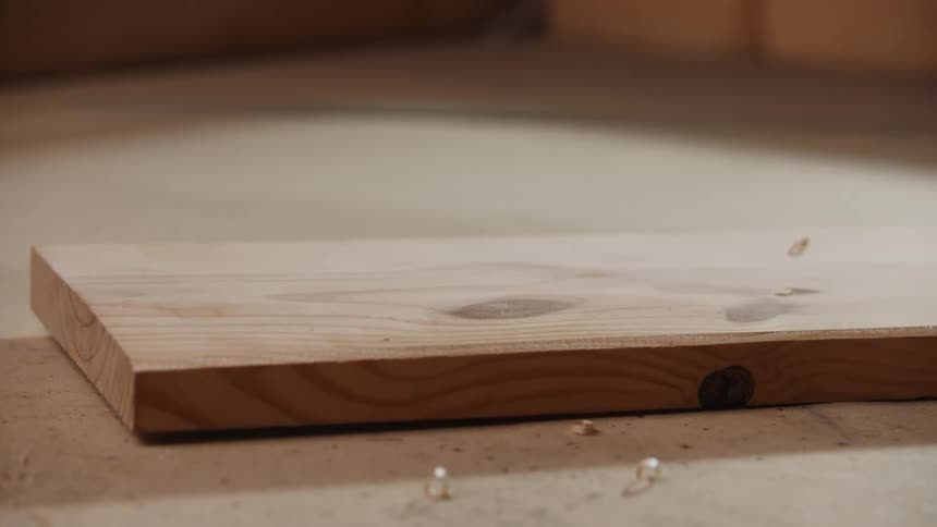 A piece of lightcolored raw wood with visible grain is resting on a workbench a hand quickly sweeps across the wood appearing to wipe away sawdust or debris