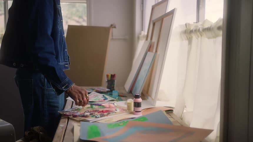 The video shows an artist in a bright studio wearing a denim jacket and jeans standing over a table covered with paint brushes and paper the artist picks up a brush and begins mixing colors or painting a small detail on the messy work surface