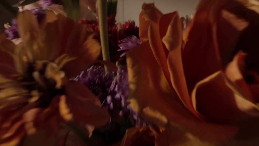 The video shows a closeup arrangement of flowers primarily featuring large orange roses and small purple flowers a persons hands are briefly visible at the beginning adjusting the flowers in the vase
