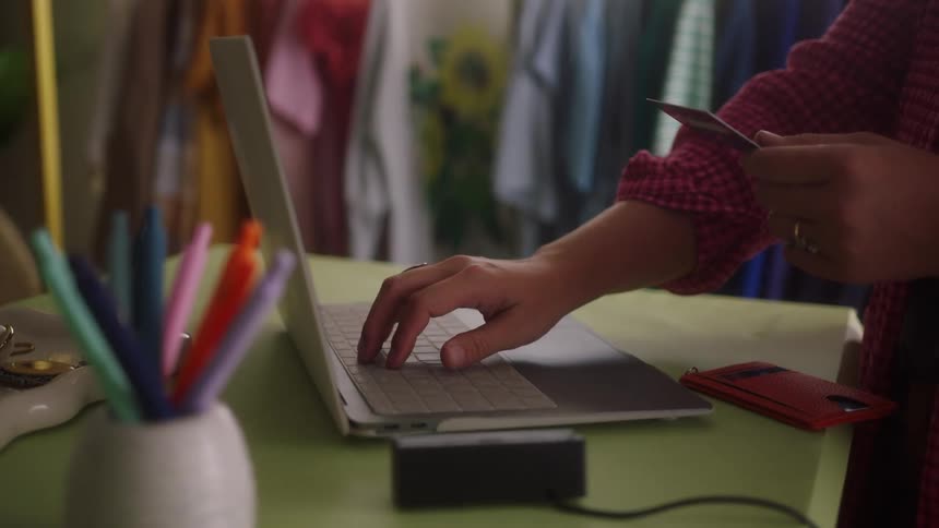 Person types on laptop while holding card over green table with red wallet and stationery items