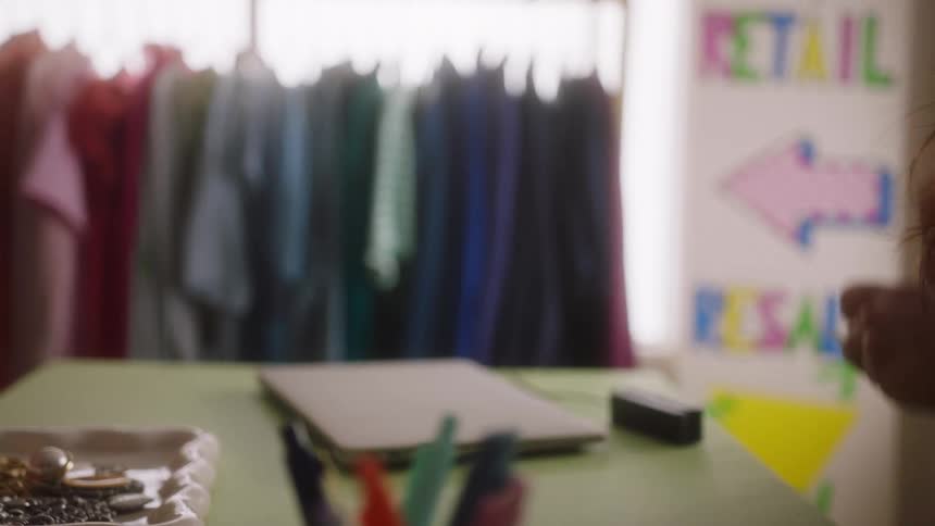 A shop clerk hands a customer a paper bag with the receipt attached completing a purchase over a counter with a rack of colorful clothing in the background
